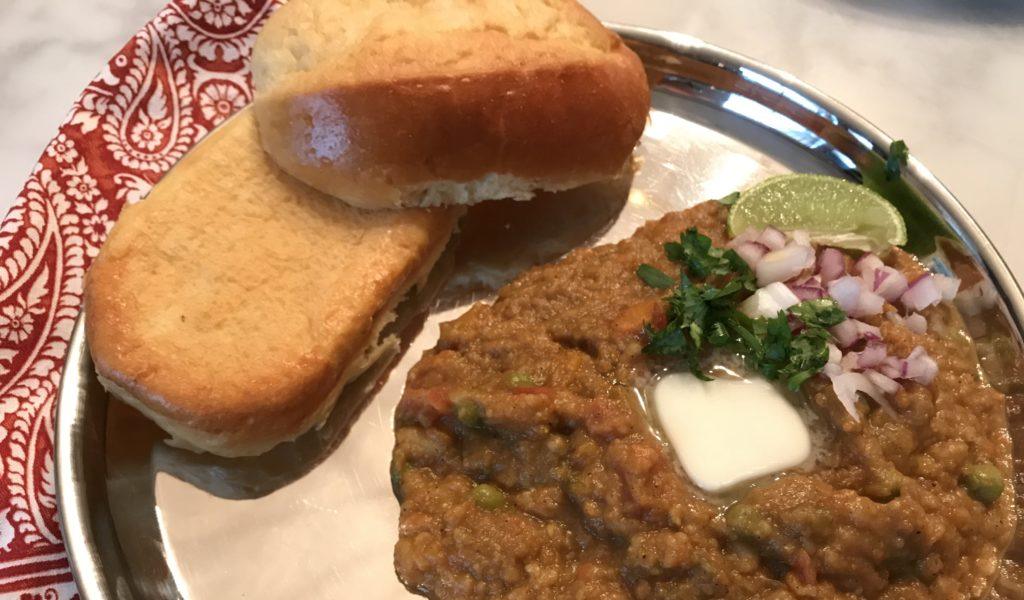 a hot plate of pau bhaji and garnished with butter, red onion, lime and cilantro and brioche rolls