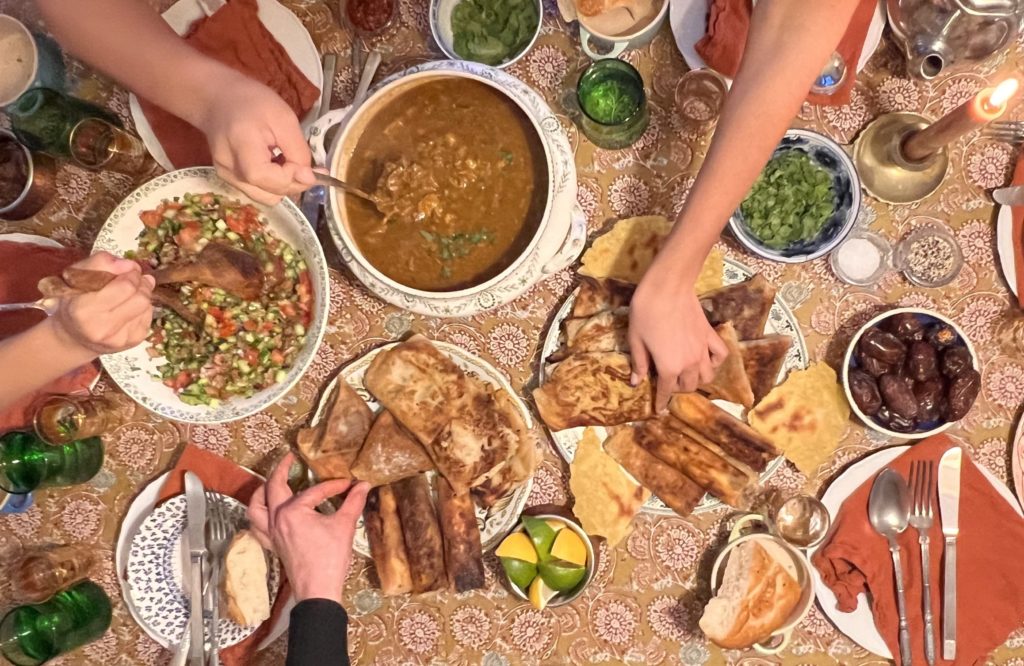 table full of homemade algerian food for soraya's algerian iftar. hands reaching for chorba, salad, bricks and bread